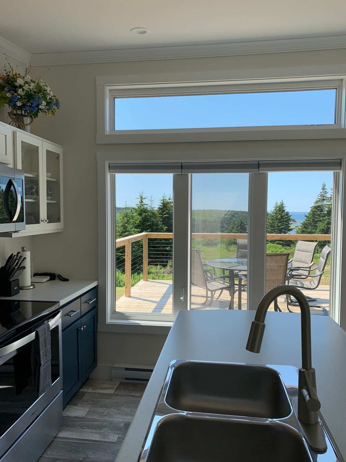 A modern kitchen is equipped with stainless-steel appliances and a double sink. Large windows provide a view of the outdoor deck and surrounding greenery, allowing natural light to illuminate the space. The design combines functionality with scenic views, enhancing the cooking experience.