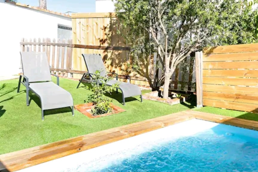 A private garden space is depicted, featuring two gray loungers positioned on vibrant green grass. A small tree is situated nearby, and a wooden fence provides privacy. A swimming pool with clear water is visible, integrated into the natural landscape.