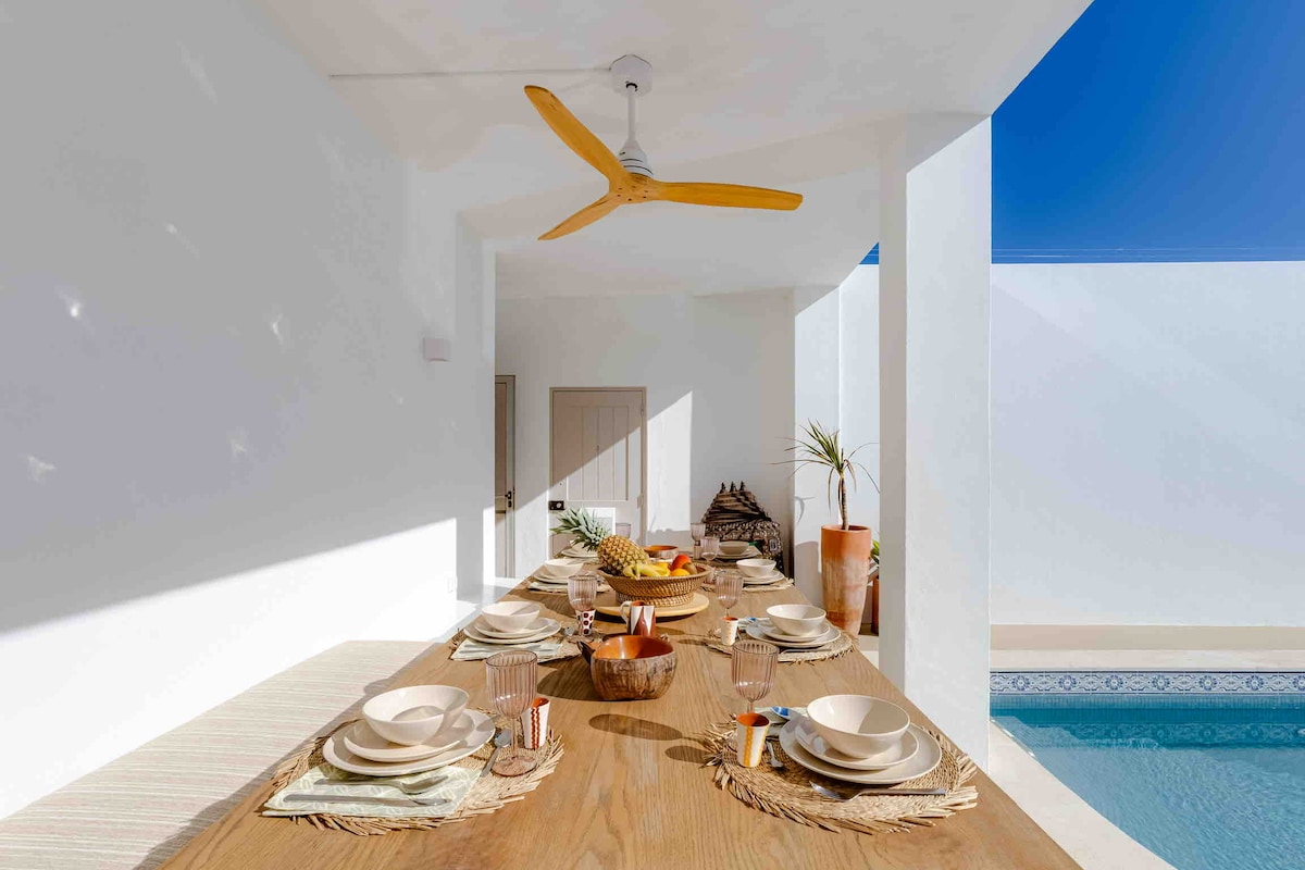 An outdoor dining area features a long wooden table set with plates and glasses. Natural light filters through spacious openings, highlighting a nearby pool and the minimalist decor of the space. A ceiling fan provides comfort under the clear blue sky.