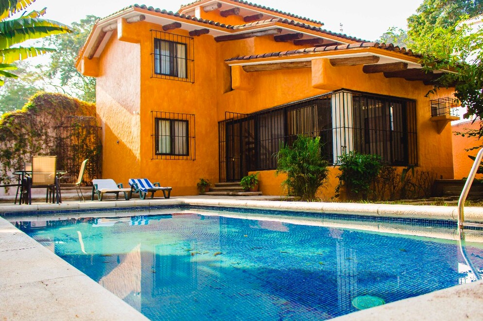 The exterior of a vibrant orange house is highlighted, featuring large windows and a tiled roof. A tranquil blue swimming pool reflects the surrounding greenery, with lounge chairs positioned nearby. Lush vegetation surrounds the property, enhancing the serene atmosphere of the home.