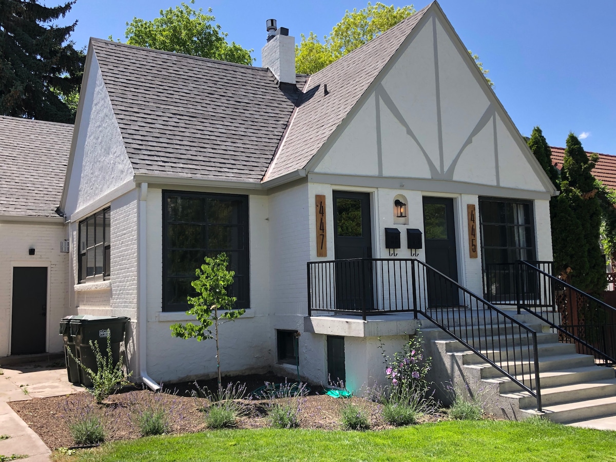 A charming exterior of a historic building showcases light-colored brick walls and a sloped roof. Large windows framed in black allow for ample natural light. A pathway leads to a set of stairs, and landscaped greenery is visible along the foundation.