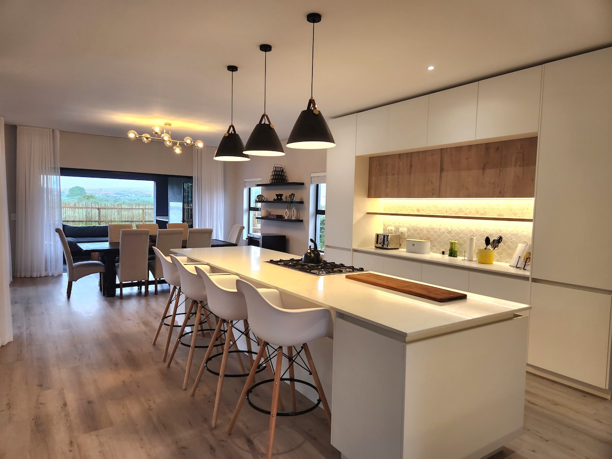 The modern kitchen features a spacious island with seating for six, complemented by sleek white cabinetry and warm wood accents. Stylish pendant lights hang above the island, illuminating the open space that flows into a dining area with large windows showcasing a scenic view.