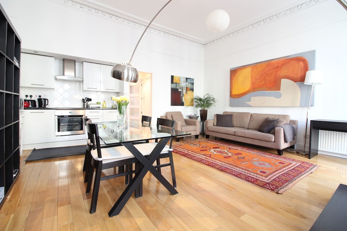 A spacious living area showcases a modern kitchen with white cabinetry and stainless steel appliances. A glass dining table is positioned near a colorful rug, while comfortable seating is arranged around a decorative wall art piece. Natural light fills the room from high windows.