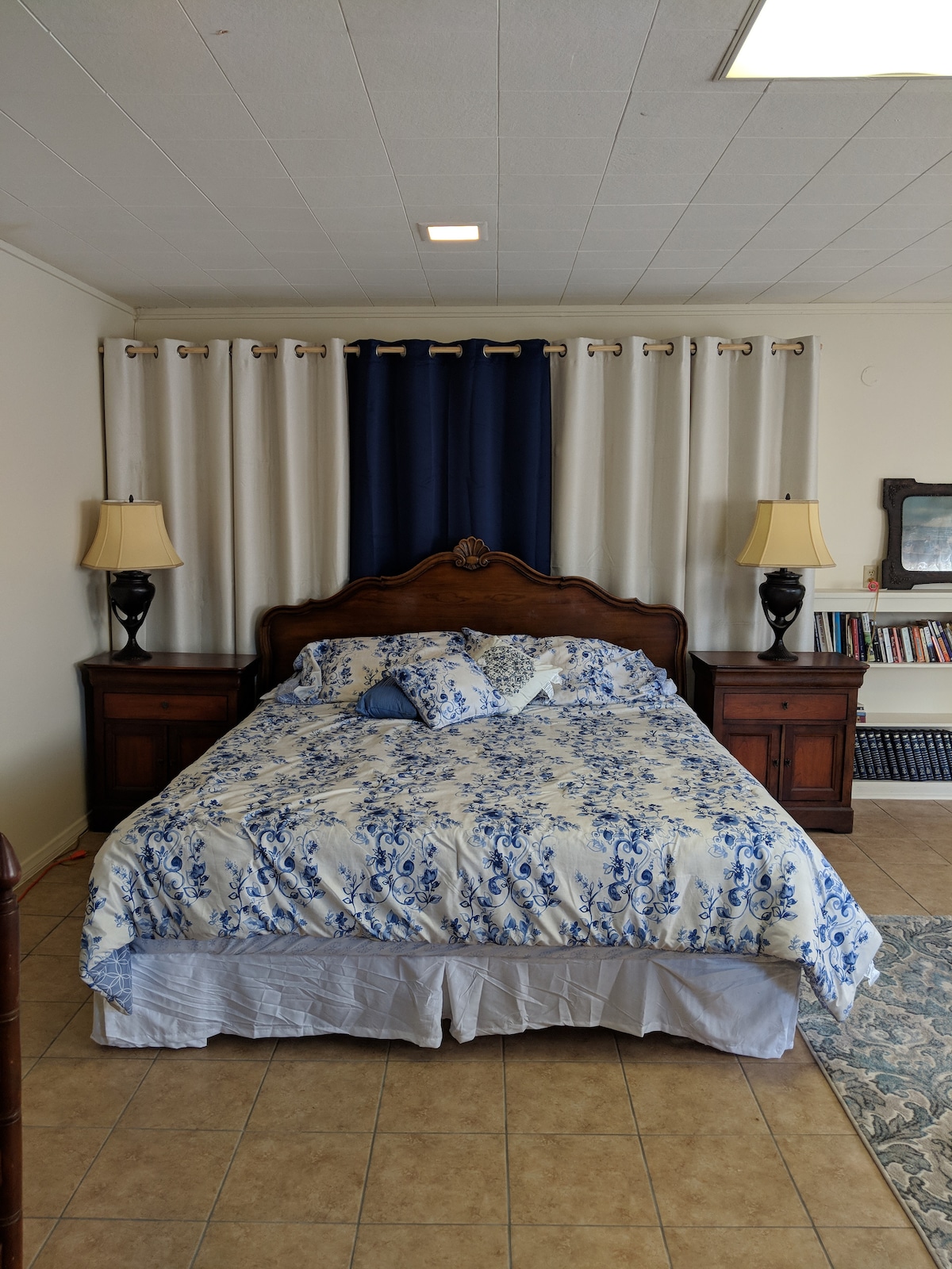 A spacious bed with a blue and white floral comforter is centered in the room, flanked by wooden nightstands with lamps. Light curtains frame a window, and a patterned rug lies on the tiled floor, creating a calming atmosphere.