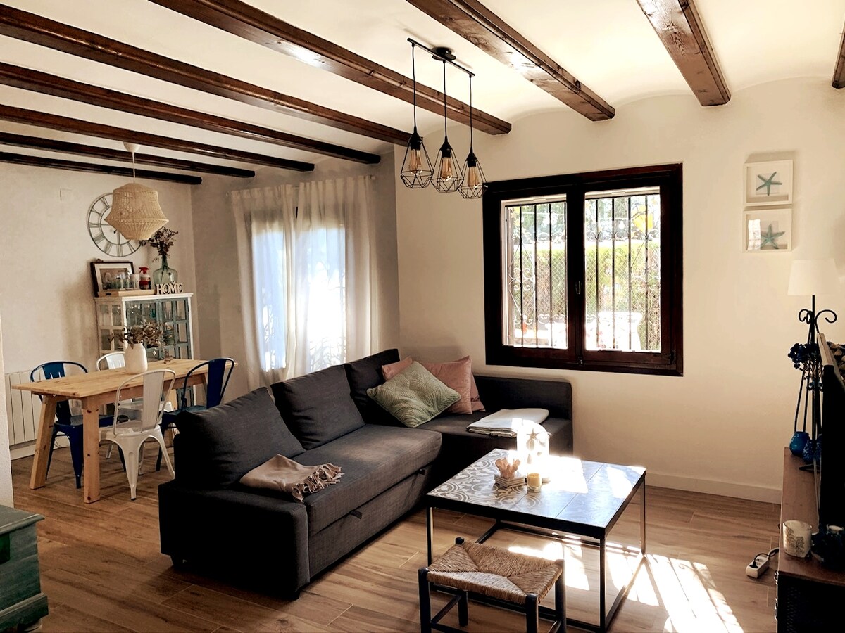 A comfortable living area features a dark sofa and a glass coffee table. Natural light streams through the window, illuminating the wooden beams overhead. A dining table with chairs is positioned nearby, alongside a decorative cabinet displaying various items.