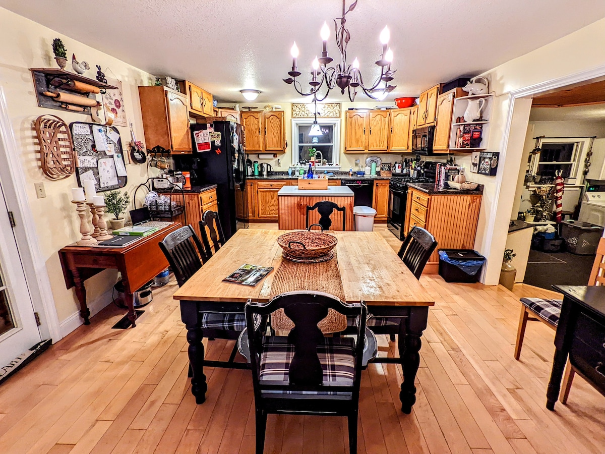 A welcoming kitchen with wooden cabinetry and a dining table at its center. The table is surrounded by dark chairs, while a chandelier hangs above. Light streams through a window, illuminating the space and highlighting the appliances and additional decor along the walls.