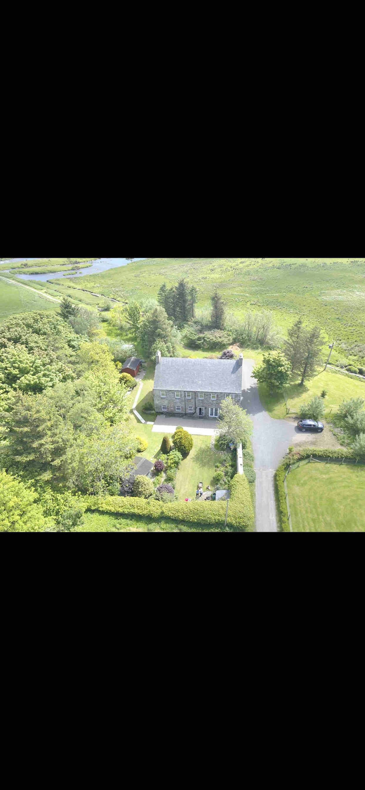 An aerial view reveals a spacious house surrounded by lush greenery, established gardens, and manicured landscapes. The building features a slate roof and is positioned along a winding path, leading to nearby natural scenery and open fields.