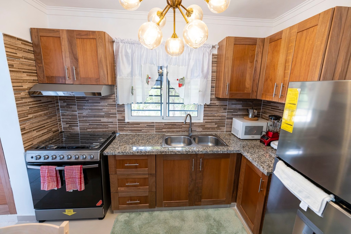 A modern kitchen features wooden cabinetry and a granite countertop. Stainless steel appliances, including a stove and microwave, are visible. A double sink is positioned under a window with light curtains. Decorative towels hang on the stove, and a round chandelier provides overhead lighting.