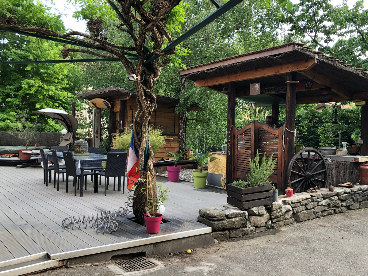 An outdoor terrace area is presented, featuring wooden structures and seating arrangements. Tables and chairs are arranged under a canopy of greenery. Potted plants in various colors adorn the space, alongside decorative elements like a vintage cart, creating a relaxed environment for guests.