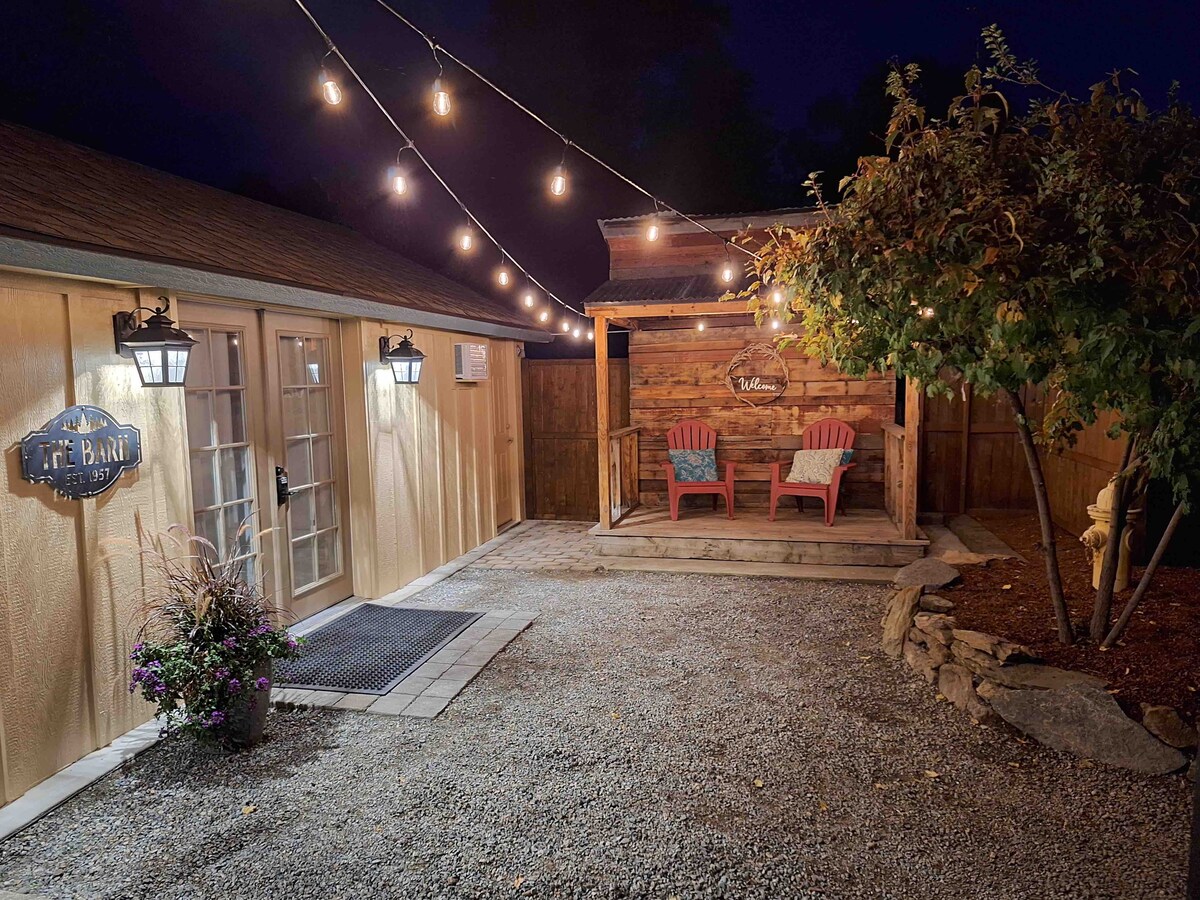 A private outdoor sitting area is featured, with two red chairs positioned under bistro lights. Gravel pathways lead to the entrance of the barn, which is adorned with lantern lighting. Lush plants are placed in decorative containers, adding natural elements to the setting.