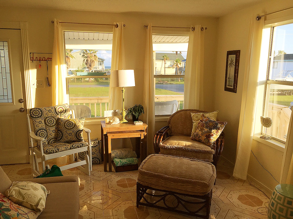 A bright living area features large windows that allow natural light to fill the space, showcasing soft yellow curtains. A comfortable wicker chair and a patterned armchair are arranged beside a small wooden table, adding a cozy touch. Decorative throw pillows and a plant enhance the inviting atmosphere.