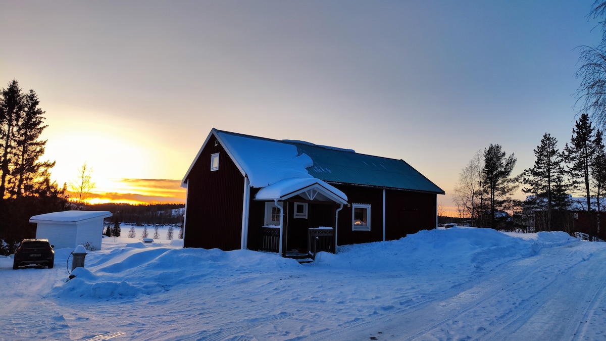 The red house, with a green roof, is framed by snow-covered ground and trees, creating a serene winter landscape. A sunset casts warm colors across the sky, contrasting with the cool tones of the snow. The driveway leads up to the entrance, inviting exploration.