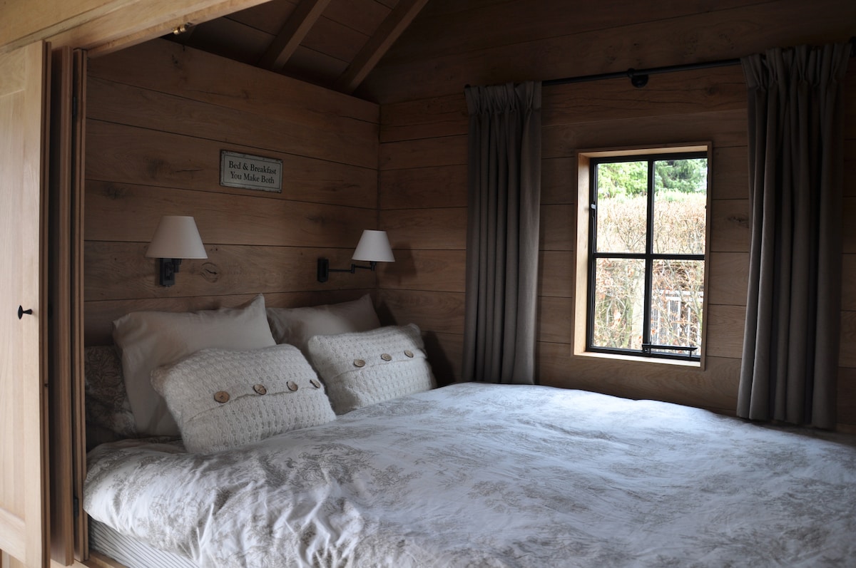 A cozy sleeping area features a spacious bed adorned with decorative pillows. Soft light enters through a window framed by simple curtains, while wooden walls add warmth to the inviting space. Wall sconces provide additional lighting for a relaxed atmosphere.