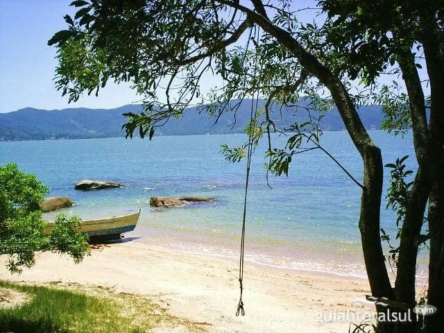 A serene beach scene is depicted with calm waters and gentle waves lapping against the shore. A small boat rests on the sand, while nearby rocks and lush greenery frame the view. In the distance, a mountainous landscape creates a peaceful backdrop.
