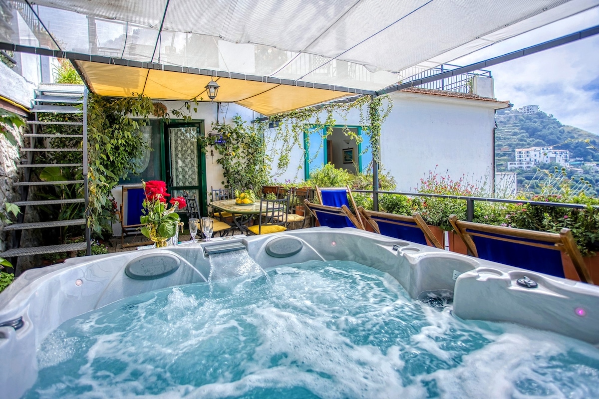 A private jacuzzi is situated on a terrace surrounded by lush greenery and vibrant flowers. Comfortable seating options are arranged nearby, creating an inviting space for relaxation. A partially shaded area provides protection from the sun, while scenic views of the Amalfi Coast are visible.
