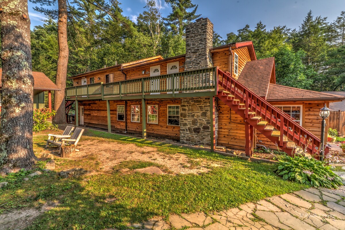 A charming wooden cottage is featured, set amidst trees in a natural landscape. A stone chimney adds character, while a balcony provides an inviting outdoor space. Steps lead up to the upper level, and a small seating area with adirondack chairs is positioned on the lawn.
