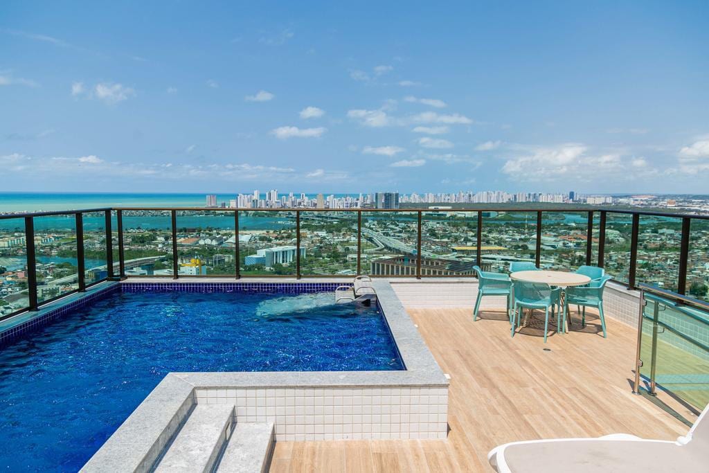 An outdoor terrace features a swimming pool with vibrant blue water, surrounded by a glass railing. Lounge chairs and a dining table with four chairs offer seating options, while a sweeping view of the skyline and ocean can be seen in the background.