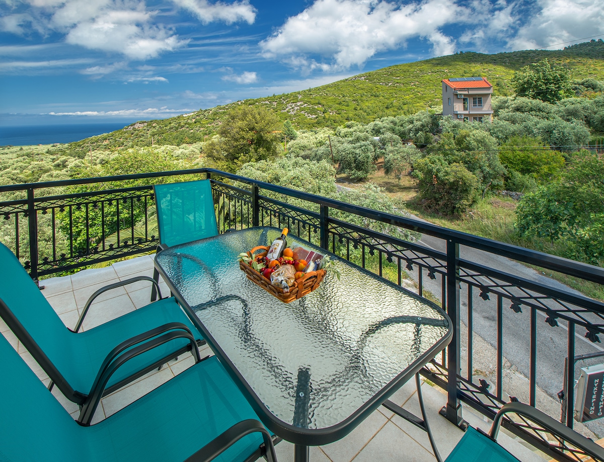 An outdoor balcony features a glass table surrounded by four turquoise lounge chairs. A basket of fruits is placed on the table. The view includes lush greenery and rolling hills, with the sea visible in the distance under a partly cloudy sky.