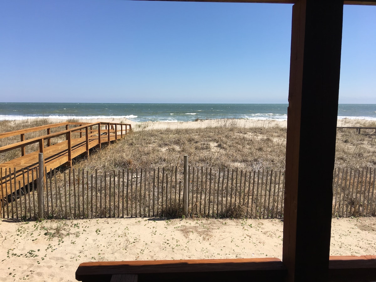 A clear view of the ocean is presented from a wooden porch, with gentle waves visible on the horizon. A sandy beach stretches below, bordered by tall grasses and a wooden walkway leading to the shore. The sky is bright and blue, enhancing the coastal scenery.