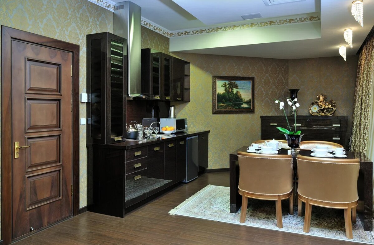 The kitchen area features dark cabinetry with modern appliances, including a refrigerator and an oven. A dining table is arranged with stylish chairs and elegantly set with tableware. Decorative artwork and a tasteful floral arrangement enhance the sophisticated atmosphere.