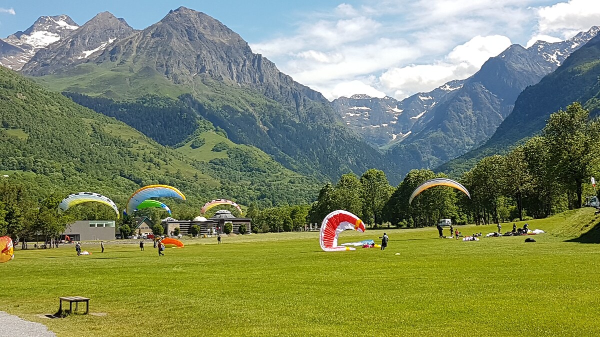 The image captures a vibrant valley surrounded by mountains, where several colorful paragliders are in the air. Lush green grass covers the ground, accommodating people engaged in various activities, with the backdrop of snow-capped peaks in a clear blue sky.