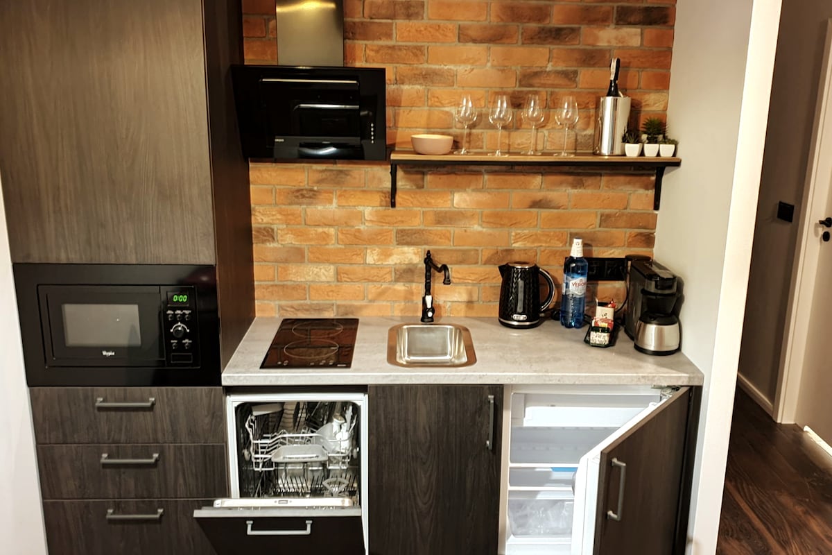 A fully equipped kitchen features dark cabinetry and a stylish brick backsplash. Included are a microwave, an induction cooktop, a sink, and a dishwasher. A coffee maker and an electric kettle are visible, alongside a shelf holding glassware and a bottle of wine. The refrigerator is open, displaying its interior.