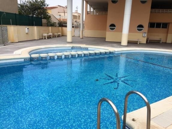 A community swimming pool features a clear blue surface and is framed by light-colored tiles. Surrounding seating areas are visible, providing space for relaxation. A gently sloped entry leads into the water, with geometric patterns visible on the pool floor.