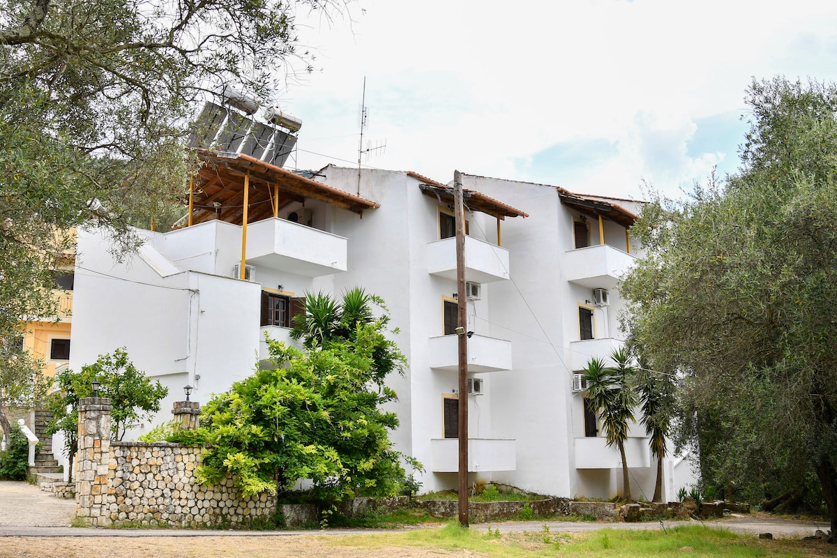 The building features a modern white exterior complemented by natural vegetation. Multiple balconies extend from the structure, surrounded by well-maintained greenery. Solar panels are visible on the roof, showcasing a commitment to sustainability. The entrance is welcoming and accessible, leading to an inviting atmosphere.