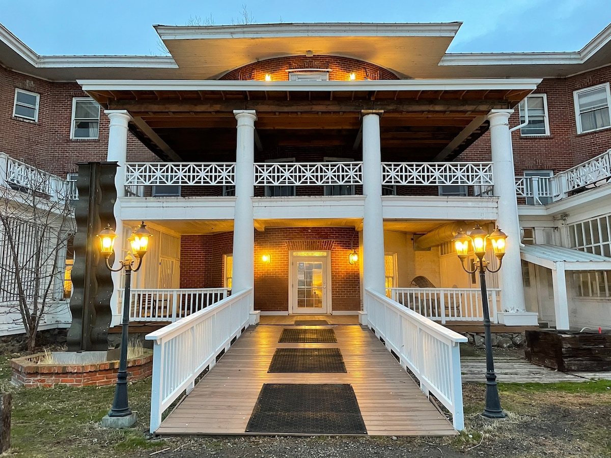 A historic brick structure features a grand entrance with large columns and a wide wooden ramp leading to the front door. Decorative lanterns illuminate the walkway, surrounded by manicured landscaping that enhances the welcoming ambiance.