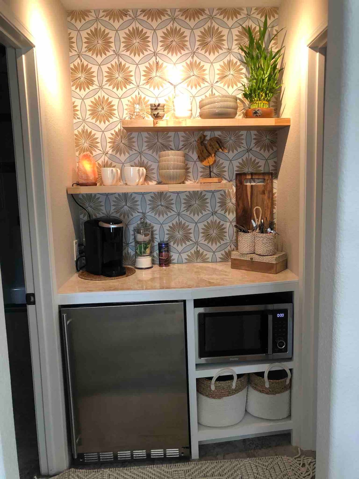 A compact kitchenette area features open wooden shelves displaying decorative items and a plant. Below, a stainless-steel refrigerator and microwave are positioned next to a coffee maker, with a selection of cups and bottles also visible on the counter.