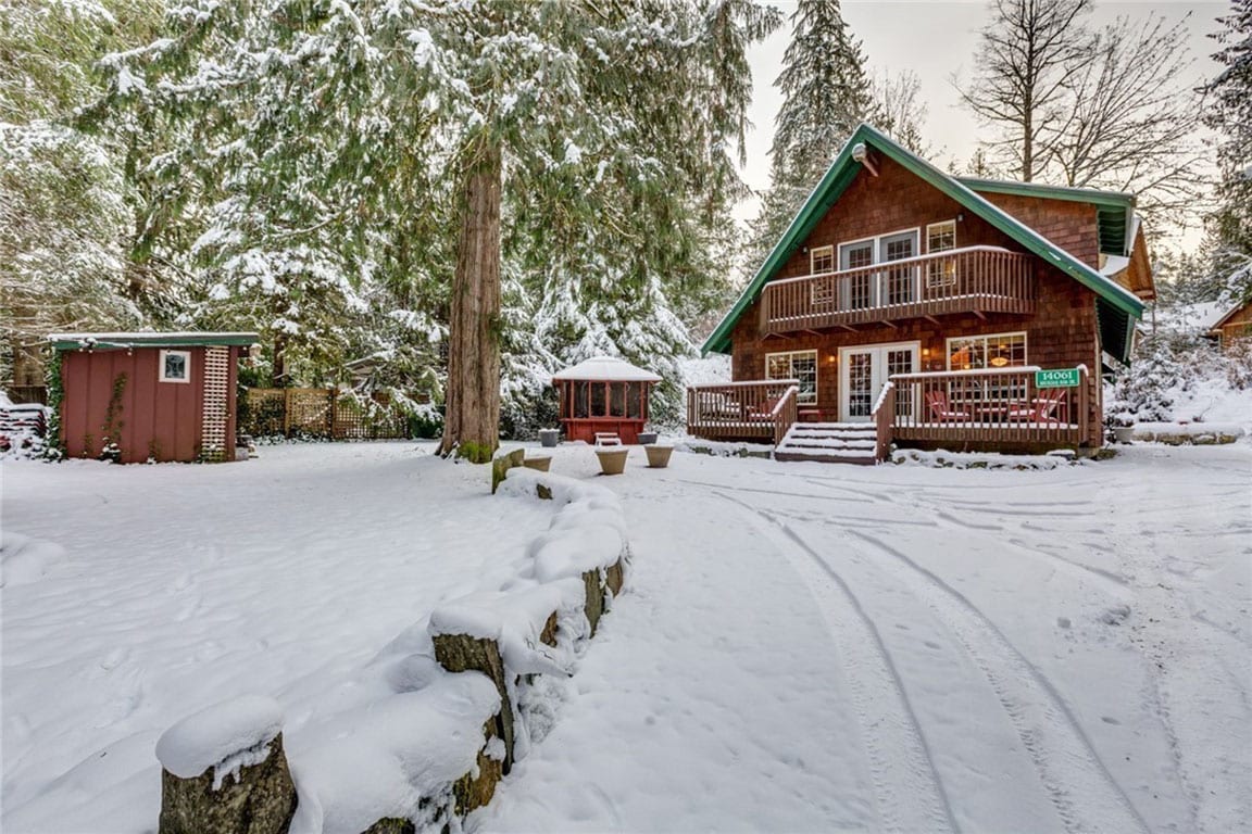 A cozy cabin is surrounded by a blanket of snow, with large trees providing a serene backdrop. Wooden decks and a gazebo are visible, inviting relaxation. A stone path leads to a fire pit area, situated in a peaceful winter landscape.