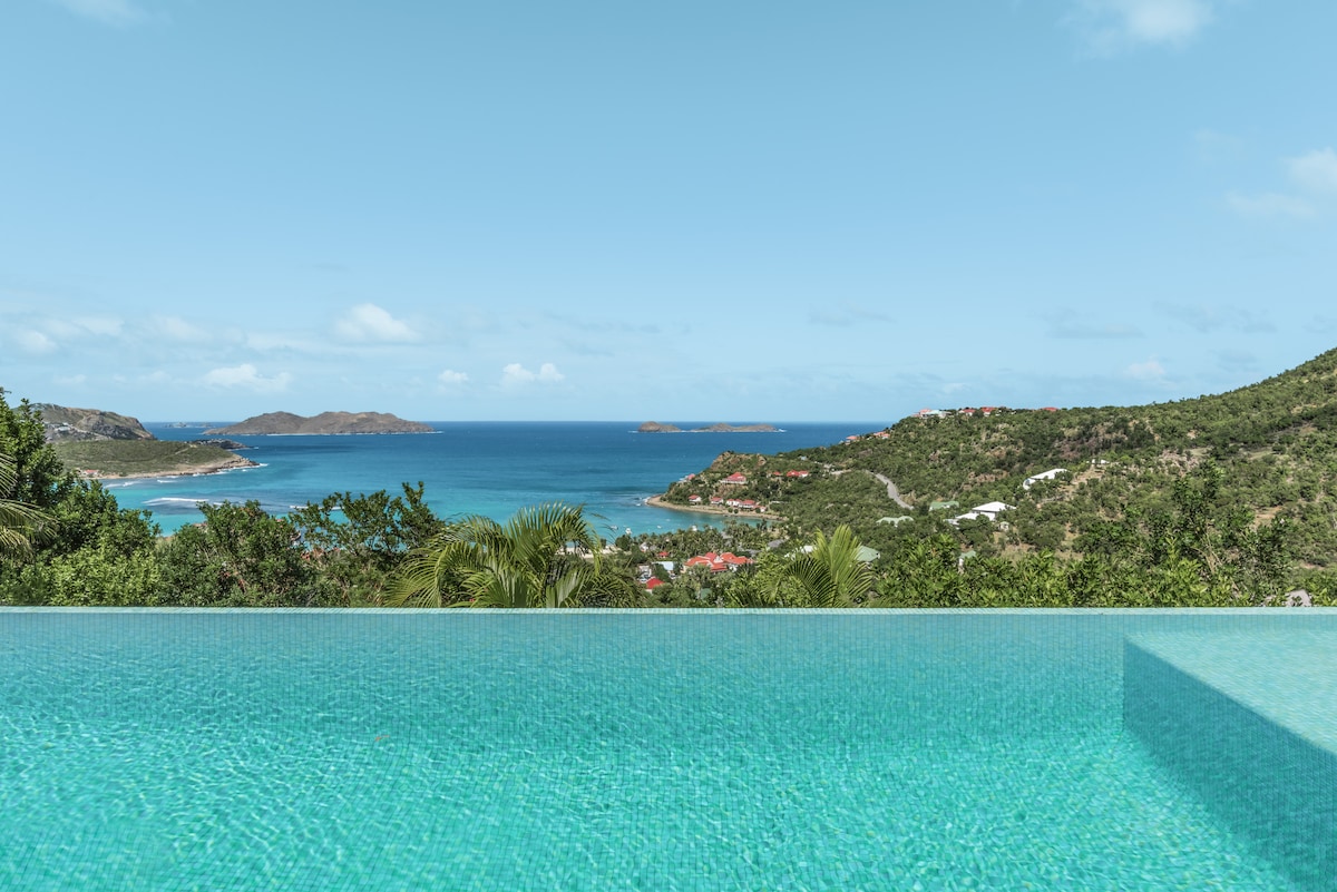 An infinity pool extends toward the horizon, blending seamlessly with the turquoise waters of the Baie de St Jean. Lush green hillsides frame the scene, while distant islands are visible in the calm sea under a clear blue sky.
