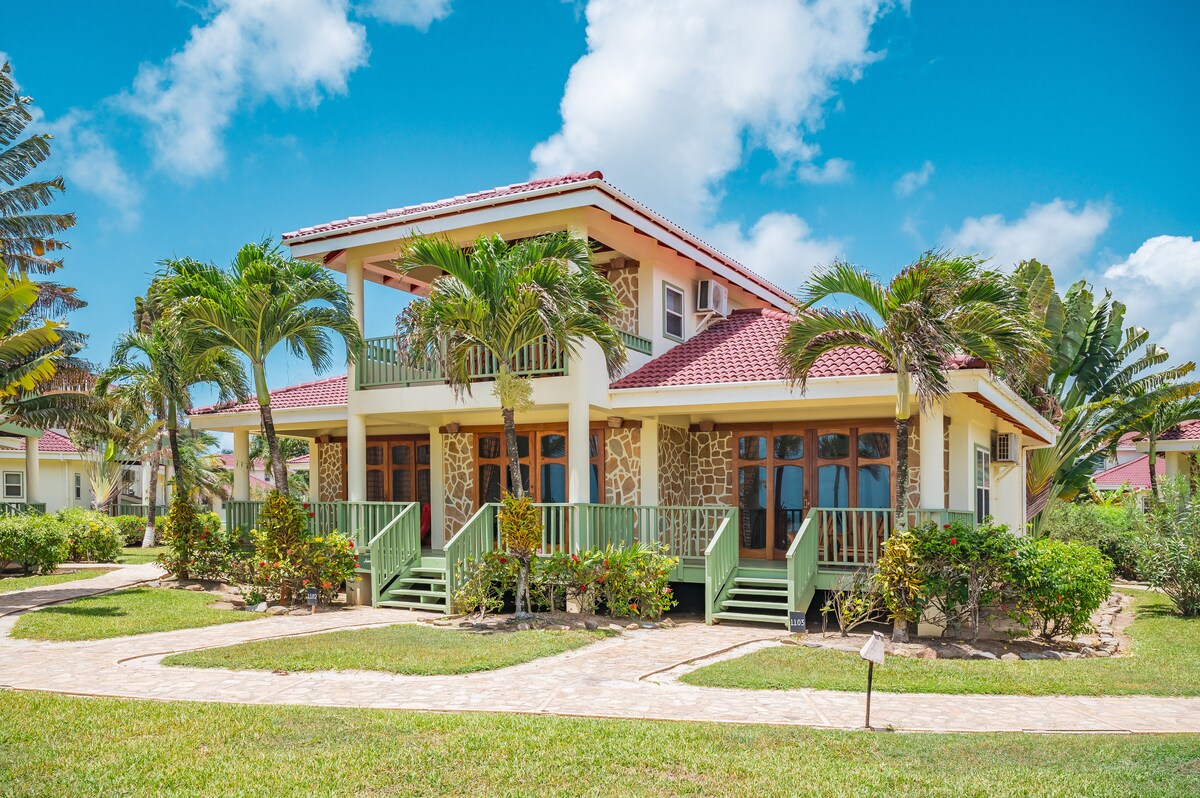 A two-story beach house is surrounded by lush greenery and palm trees, featuring a vibrant red roof. Wide wooden steps lead to the entrance, and multiple balconies are visible. The exterior showcases a blend of stone and siding, set against a clear blue sky.