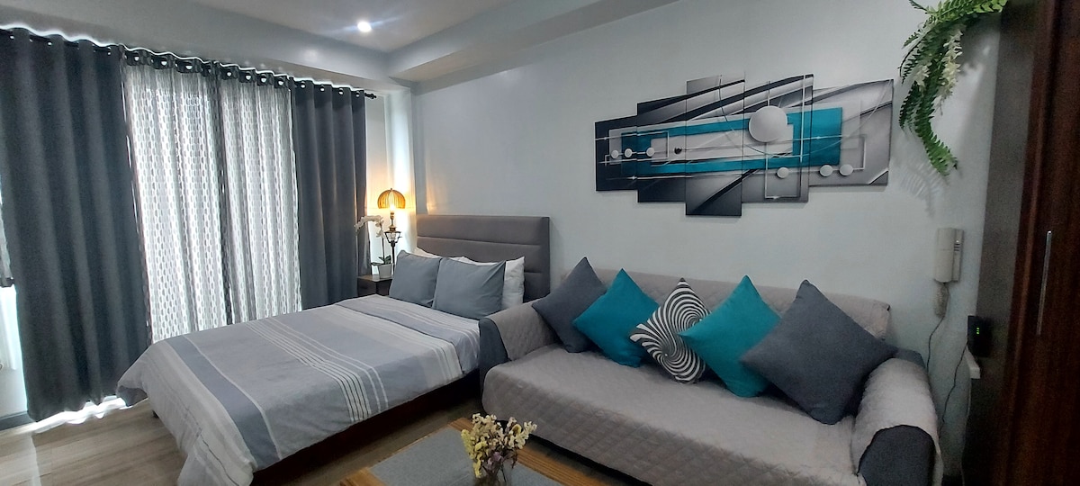 An air-conditioned studio unit features a spacious bed and a sofa. Decorative cushions in shades of blue and gray are arranged on the sofa. Natural light is allowed in through floor-to-ceiling windows covered with sheer and blackout curtains. A contemporary artwork hangs above the bed.