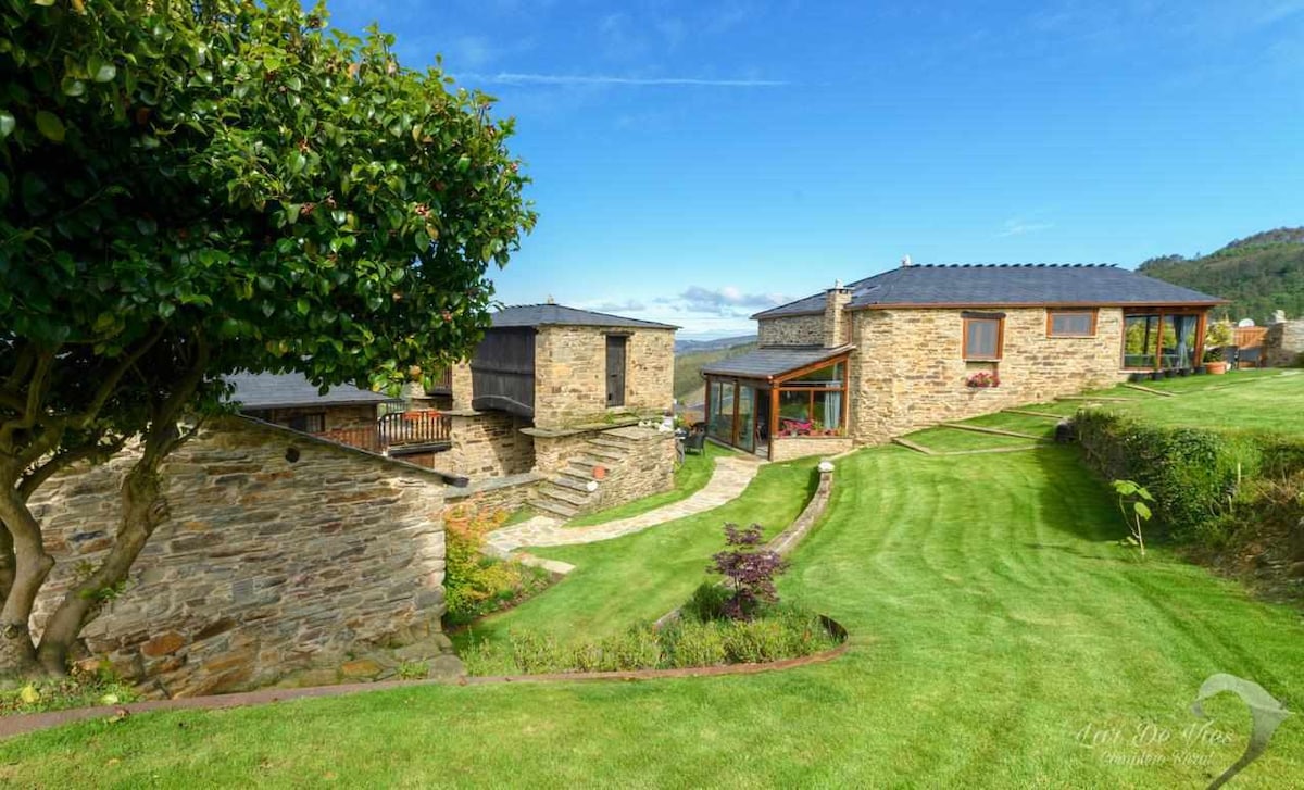 The image features a scenic view of a rustic stone complex, surrounded by well-maintained green lawns and gardens. The buildings exhibit a harmonious blend of natural materials and modern design, with a pathway leading through the lush landscape towards the structures.
