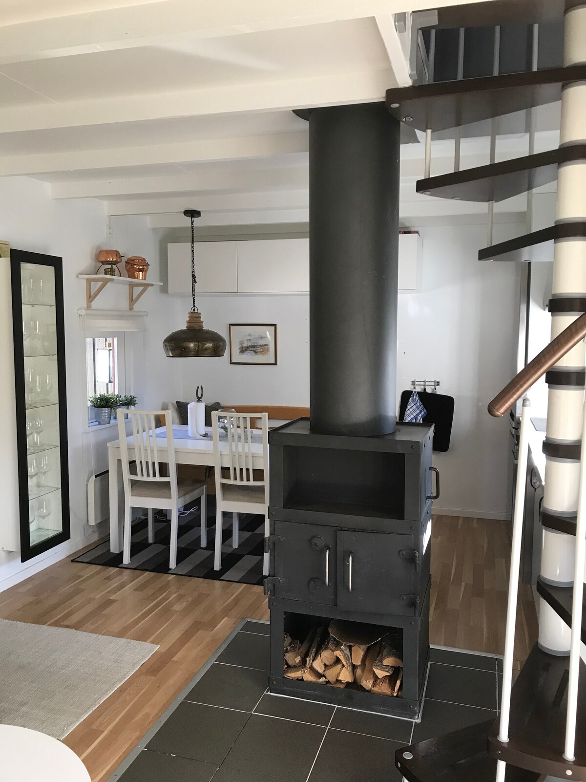 A stylish living area features a wood-burning stove, providing a focal point. A dining table with four chairs is positioned near the stove, while light wood flooring complements the space. A spiral staircase leads to the upper level, flanked by a decorative cabinet and wall art.