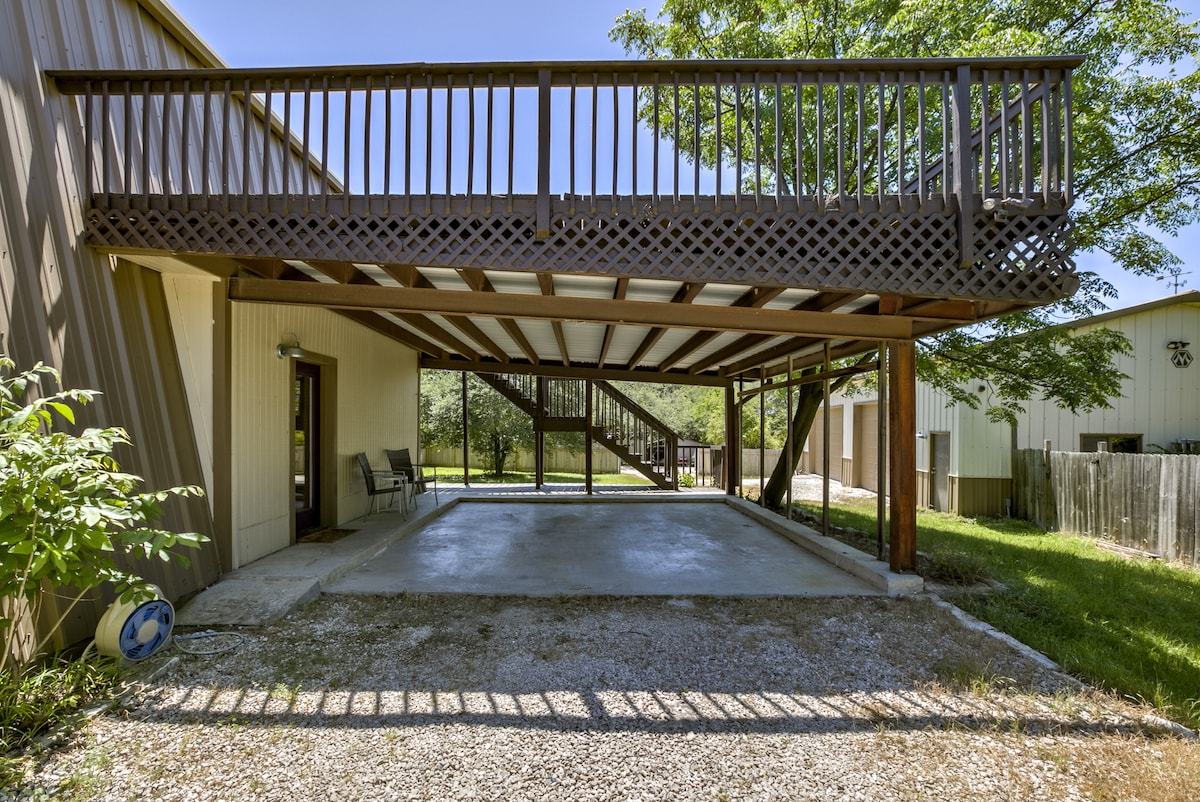 An outdoor parking area is featured under a covered wooden structure, providing shaded space. The area is surrounded by greenery, and a few chairs are placed against the wall for seating. The adjacent staircase leads to the upper level of the building.