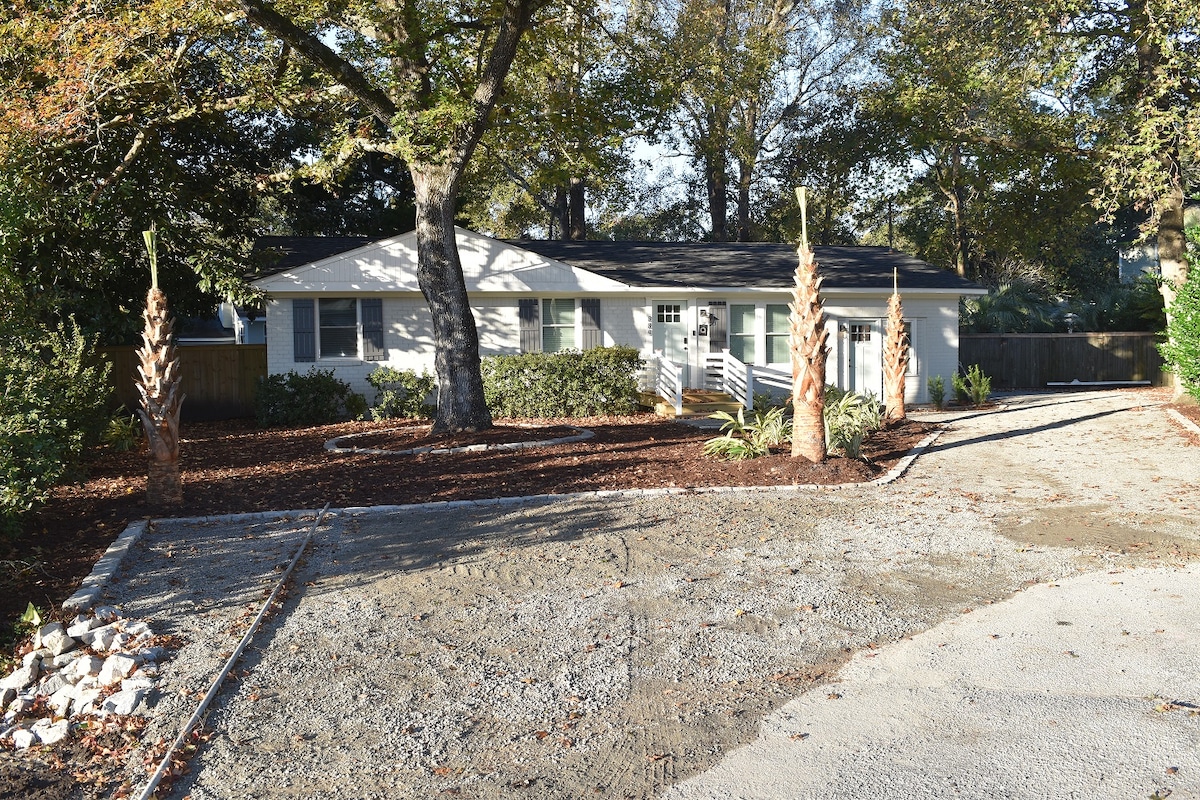 A single-story home is set among mature trees, featuring a light-colored exterior. The driveway leads to the entrance, surrounded by landscaped areas with decorative plants. The fenced yard is visible in the background, creating a sense of privacy.