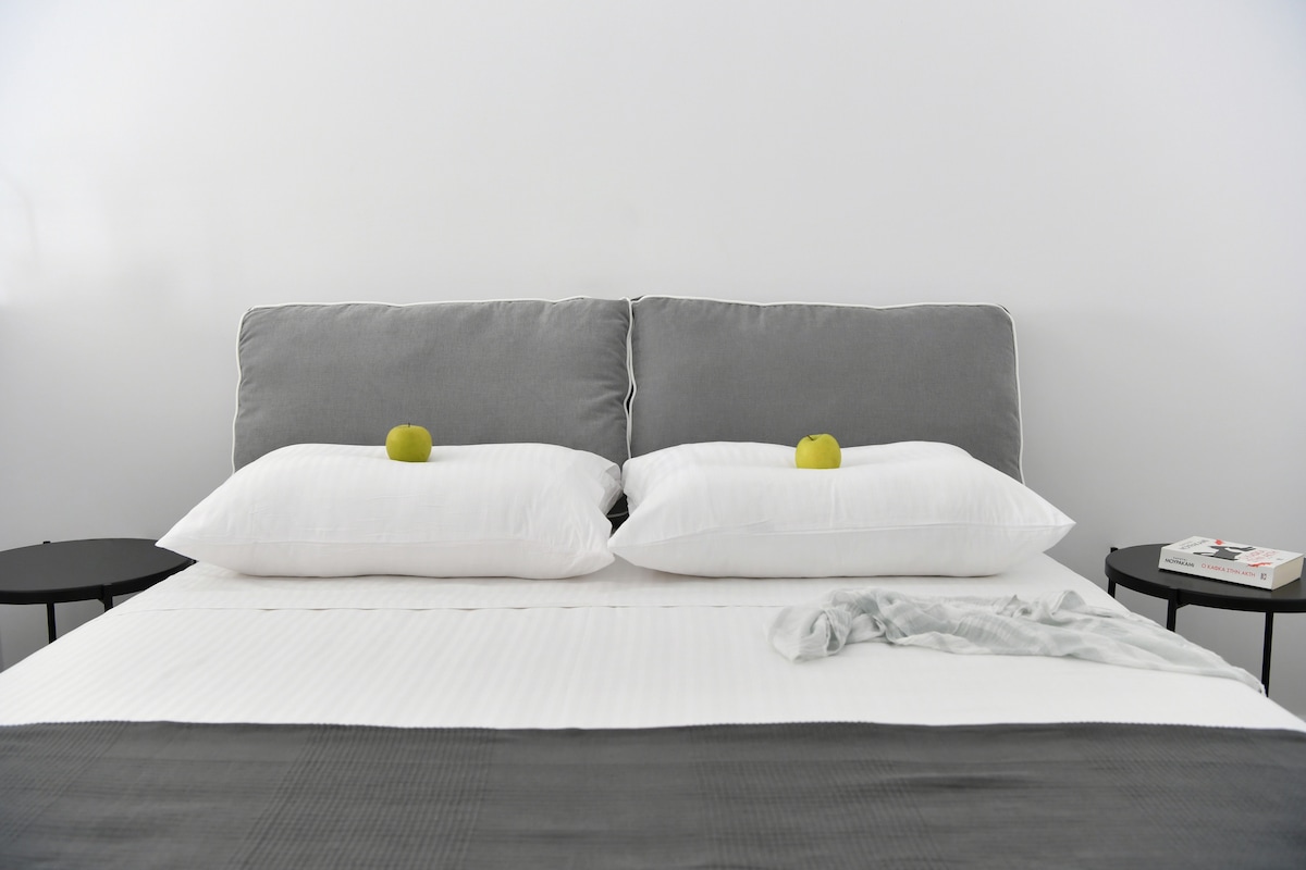 A modern bed with a gray upholstered headboard is positioned against a white wall. Two white pillows are placed on the bed, with green apples resting on top. On either side, sleek black bedside tables hold a small book and a decorative item.
