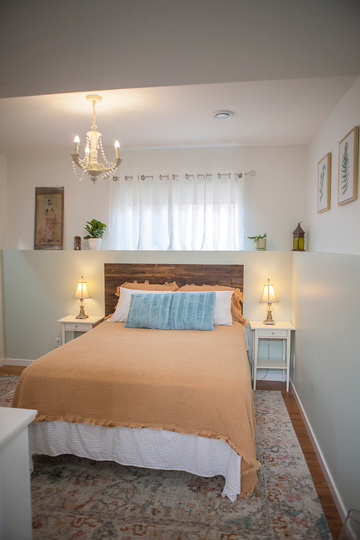 A cozy bedroom features a warm-toned bedspread draped over a spacious bed, with light streaming through sheer curtains. Two bedside tables with lamps flank the bed, while a vase with greenery adds a touch of nature to the decor.