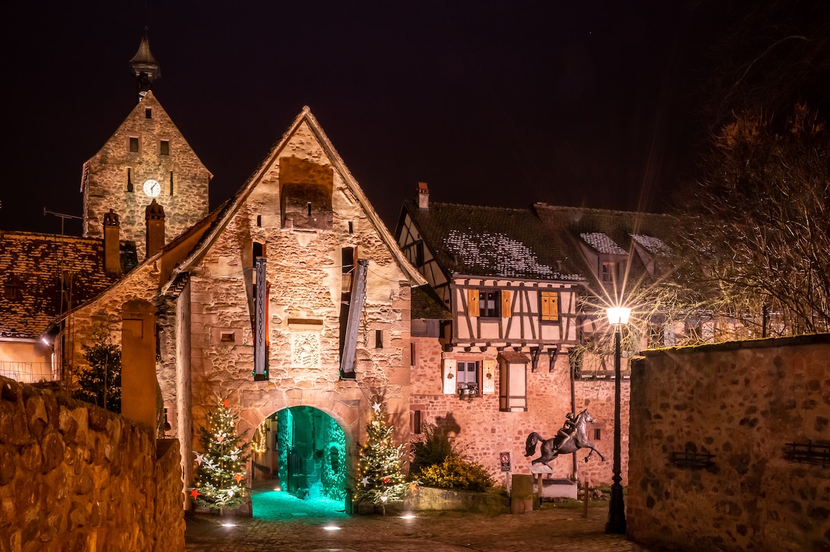 The historic building is illuminated at night, showcasing a stone façade with timber accents. Seasonal decorations, including small evergreen trees, are placed along the walkway. A softly glowing lantern provides warm light, creating an inviting atmosphere in this charming village setting.