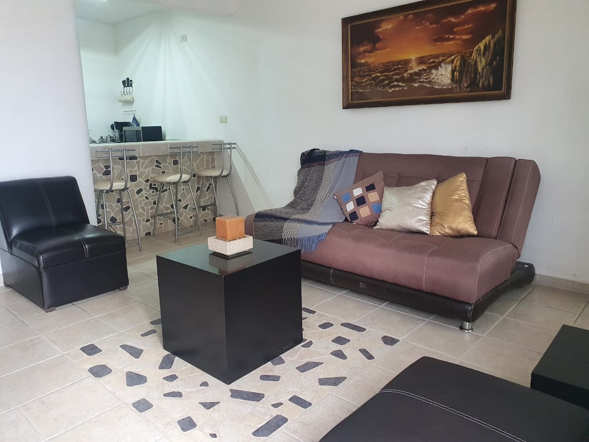 A living area features a brown sofa with decorative cushions and a sleek black coffee table set on a tiled floor. A light-colored bar area is visible in the background, complementing the overall layout.