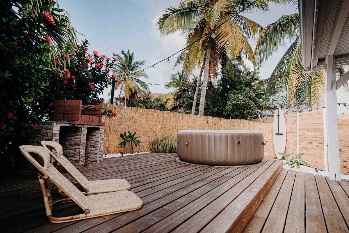 A serene outdoor space is featured, showcasing a hot tub surrounded by lush greenery and palm trees. Two woven lounge chairs are positioned on a wooden deck, offering a peaceful spot for relaxation. String lights are visible, adding a cozy ambiance to the area.