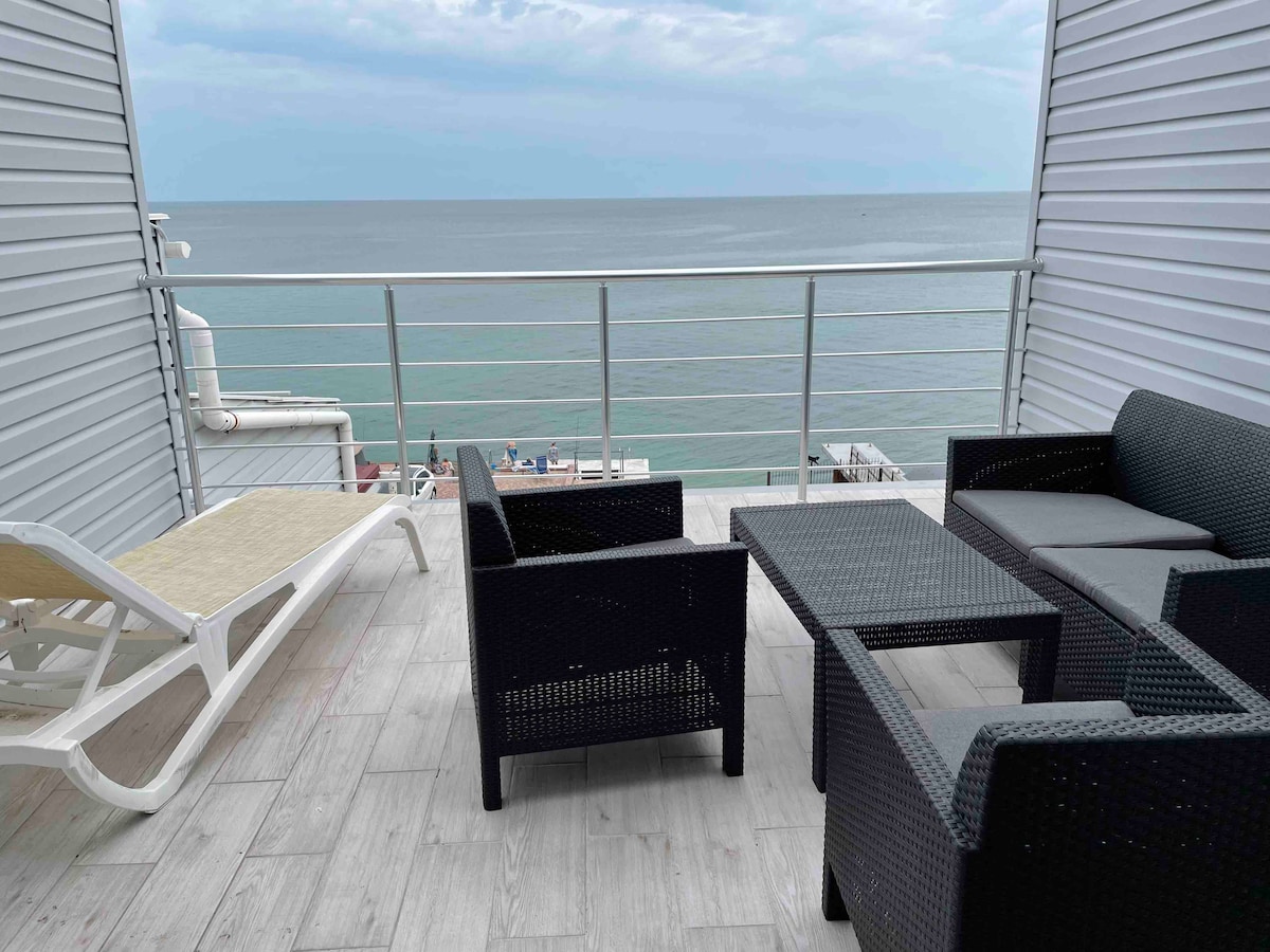 A balcony area features a dark wicker seating arrangement with a coffee table, complemented by a sun lounger. The space offers unobstructed views of the sea, creating a serene setting. The flooring is light, enhancing the overall ambiance of the outdoor space.