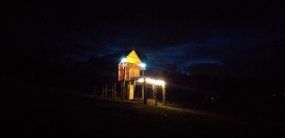 A wooden structure is visible at night, partially illuminated by soft outdoor lighting. The building features a sharply peaked roof and stands elevated on a hillside, surrounded by dark landscapes under a night sky, enhancing the sense of tranquility and seclusion.