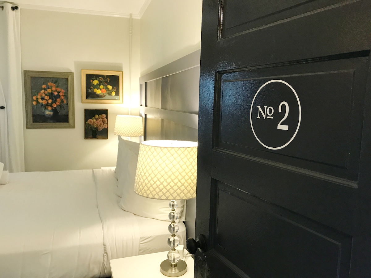 A softly lit room is framed by pastel walls, featuring a king-sized bed adorned with crisp white linens. Decorative wall art adds a touch of color. An elegant bedside lamp stands on a nightstand, highlighting the welcoming atmosphere as the door is partially open to reveal the room.