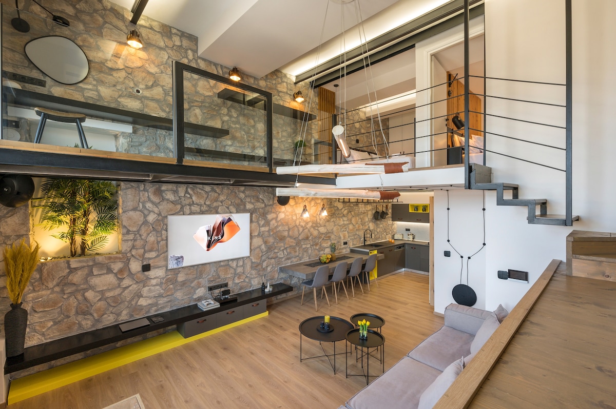 An open-concept living space features a combination of stone and wooden elements. Large windows allow natural light to fill the room, highlighting the modern furnishings, including a comfortable sofa and a dining area. A staircase leads to the upper level, providing additional living space.