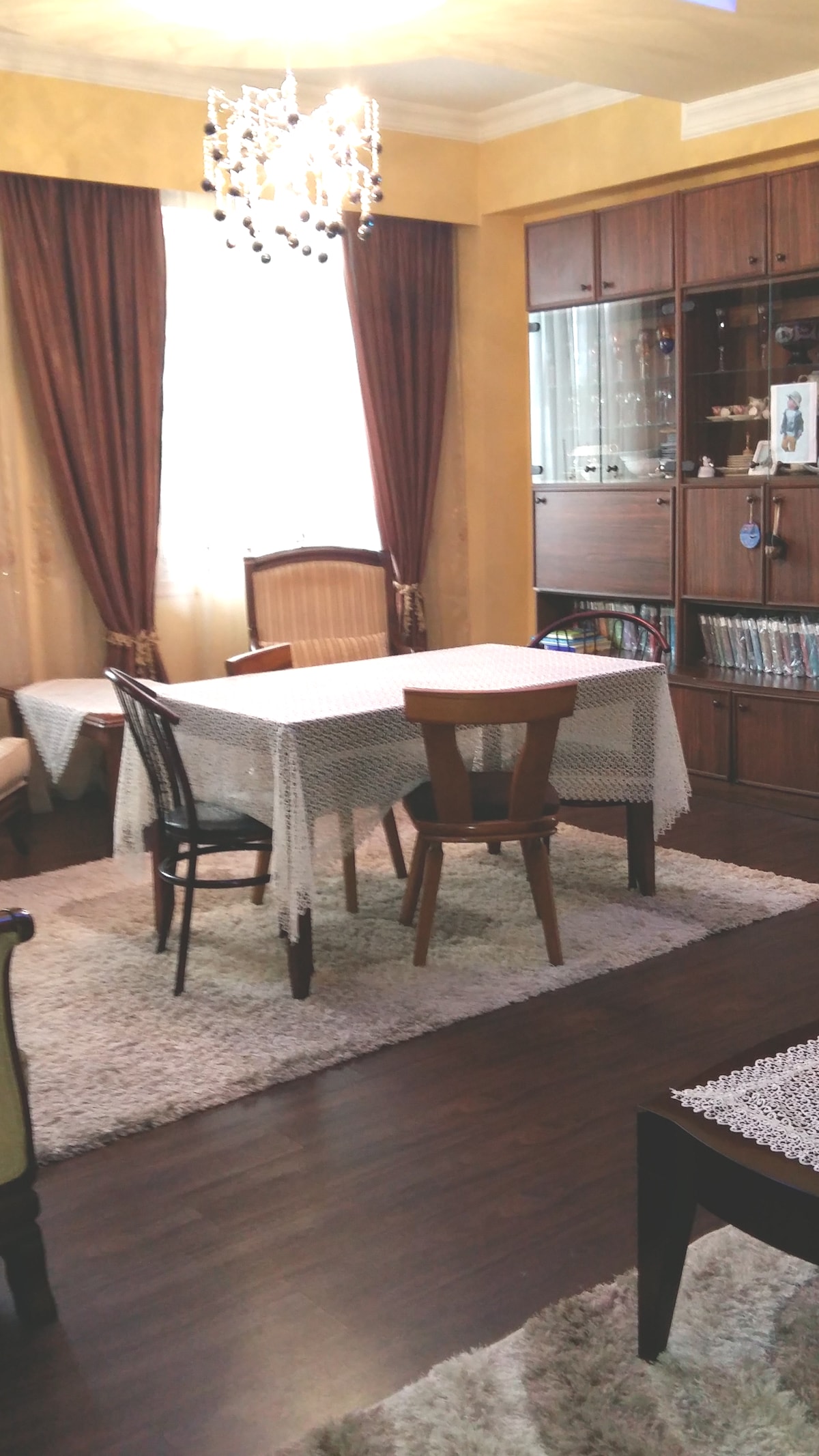 A dining area features a rectangular table with light-colored tablecloths, surrounded by a mix of chairs. Two large windows are dressed with long, flowing brown curtains, allowing natural light to illuminate the space. A rug covers part of the wooden floor, adding warmth to the room.
