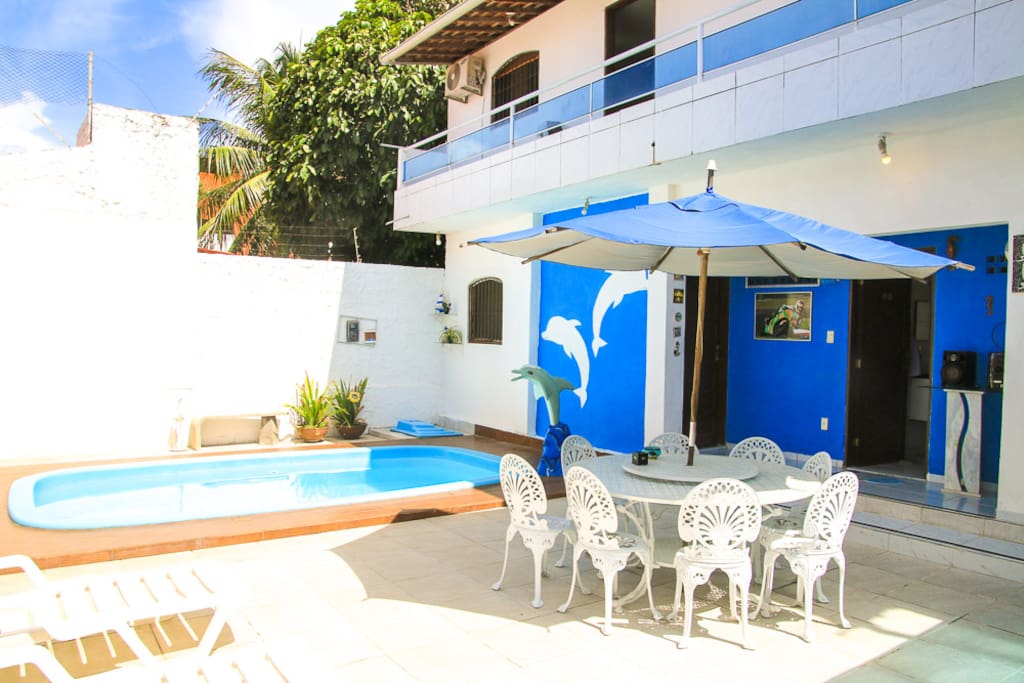 Área externa com piscina, churrasqueira, mesa, cadeiras, espreguiçadeiras e geladeiras disponíveis.