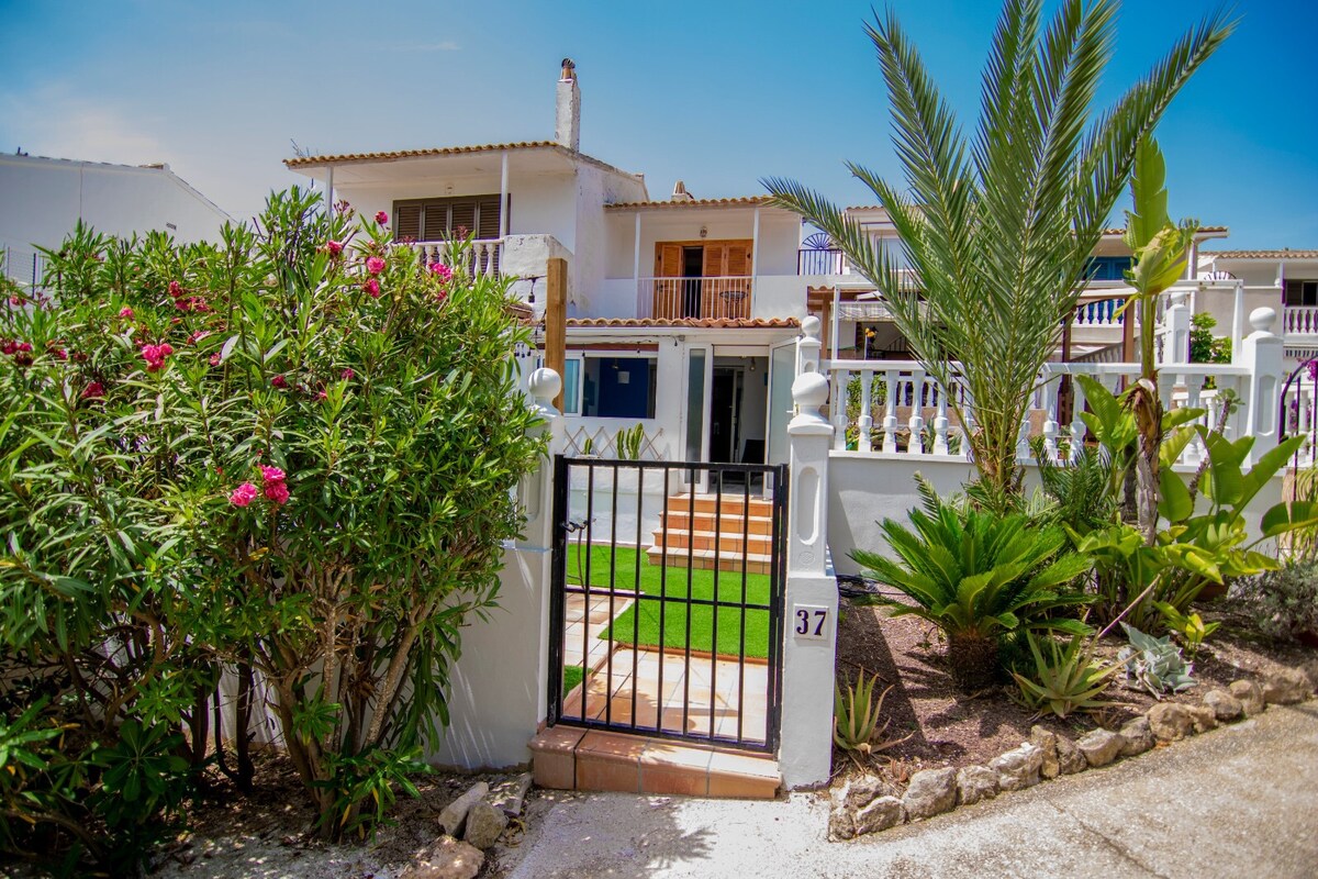 A well-maintained entrance is framed by vibrant greenery and tropical plants. The pathway, lined with decorative stones, leads to a charming bungalow with a staircase, while a clean white wall and a wrought-iron gate provide added privacy and security.